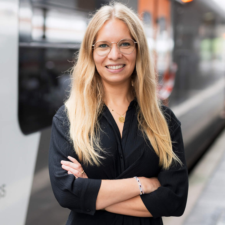 Eine blonde Frau lächelt in die Kamera. Sie steht vor einem Zug an einem Gleis.