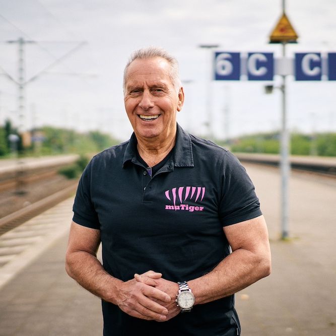 Ein Mann in schwarzem Poloshirt steht lachend am Bahnhof.